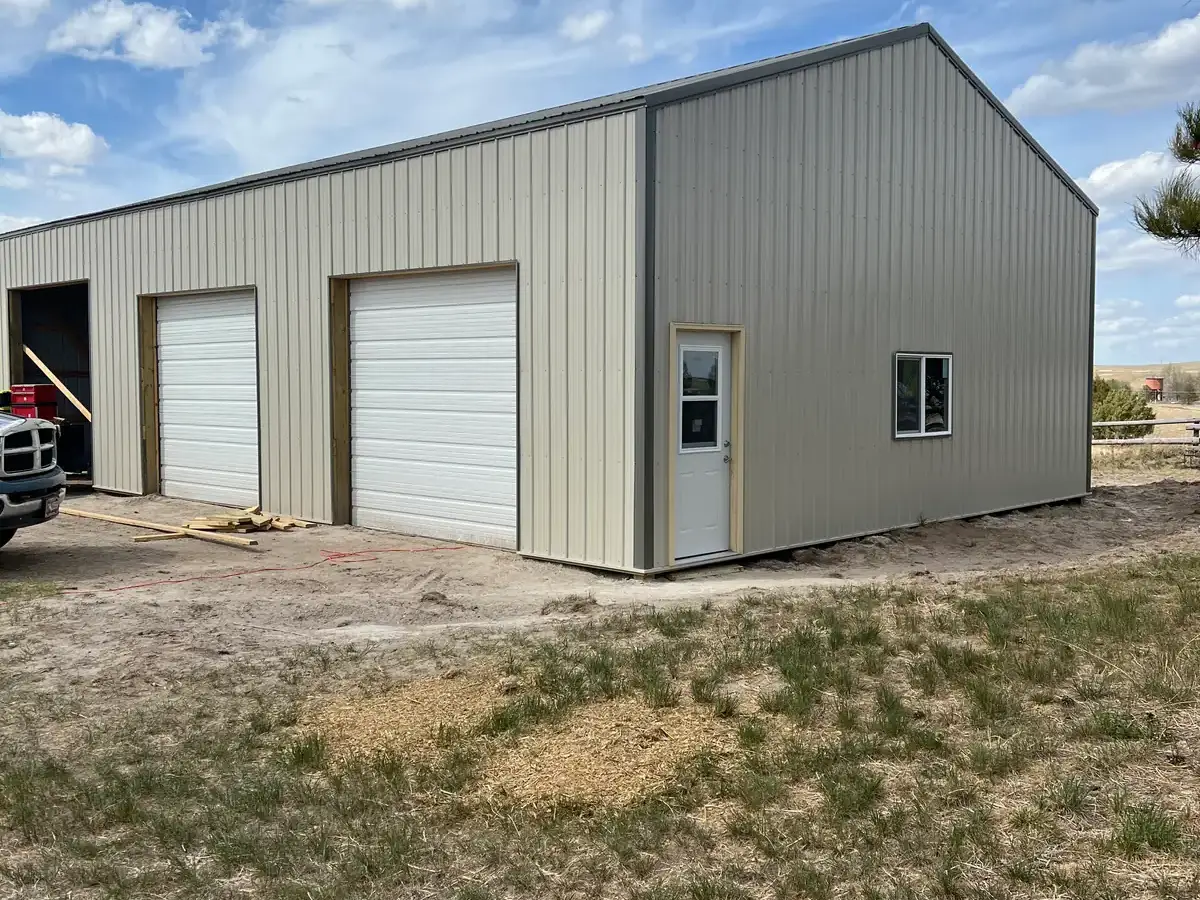 Corner view of a metal garage with white overhead doors, a side entry door and two windows.