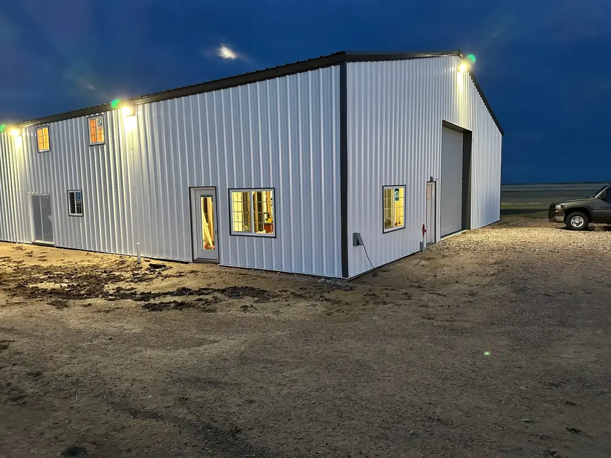 White metal building with illuminated windows, exterior lights and a large overhead door at dusk.